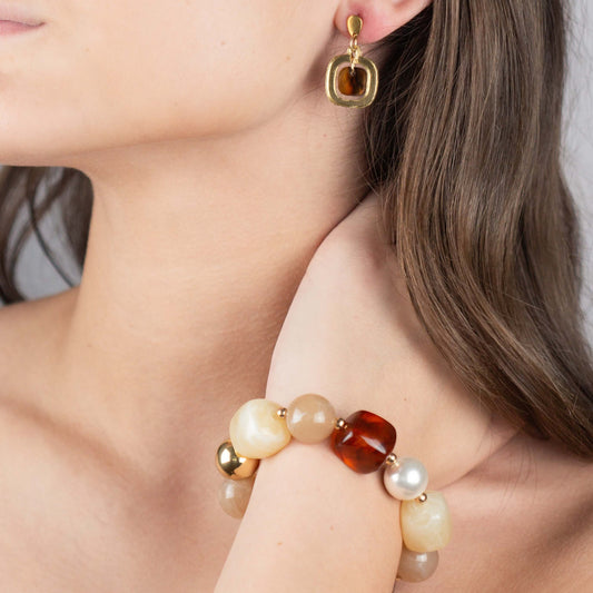 A close-up lifestyle shot of a model wearing a multi-colored beaded bracelet on her wrist and a matching gold drop earring with a brown stone.