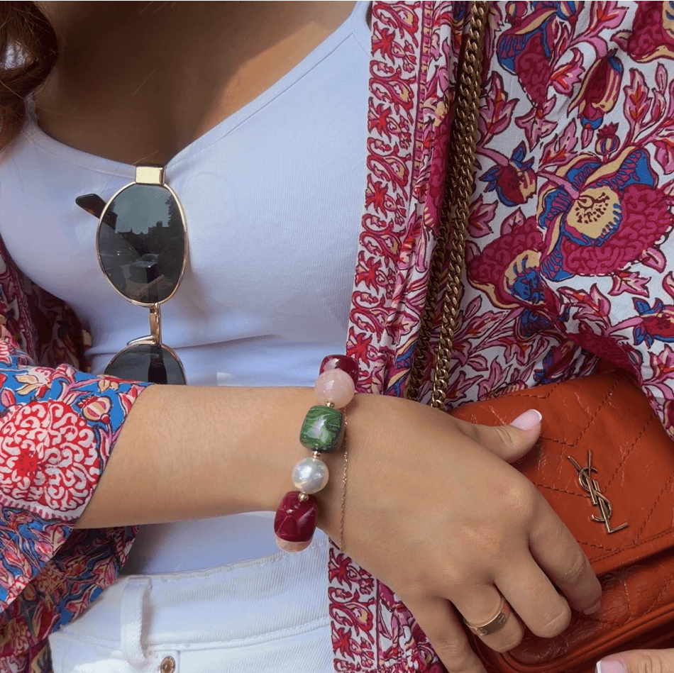 A close-up of a person's hand wearing a beaded bracelet with pink, green, and red beads, styled with a white top, sunglasses, an Yves Saint Laurent bag and a red patterned jacket.