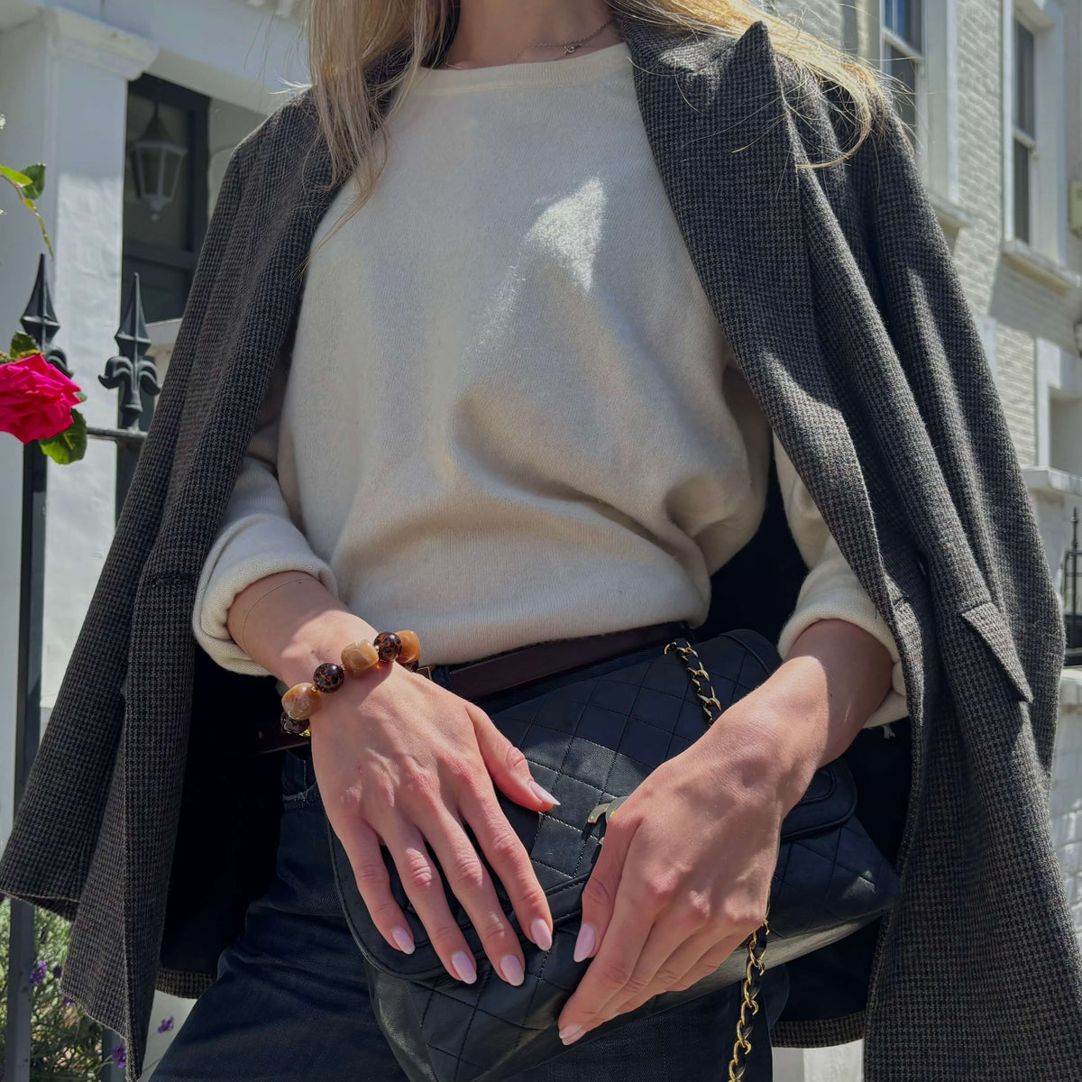 A woman wearing a cream knit sweater under an open dark gray houndstooth blazer, holding a black quilted handbag with a gold chain strap. She is wearing a brown beaded bracelet on her wrist.