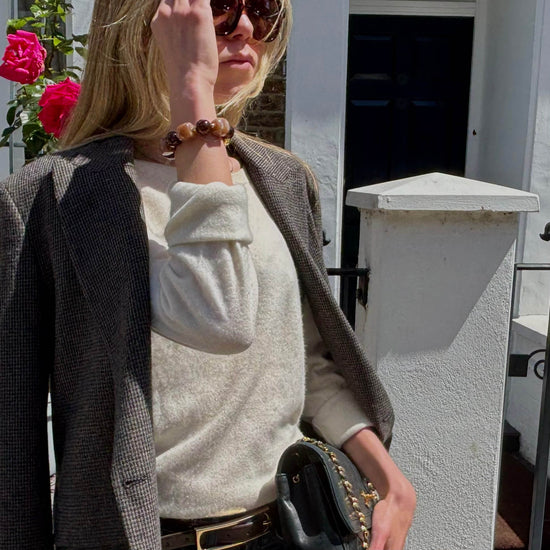 A woman outdoors wearing a cream knit sweater under a dark gray textured blazer, accessorized with a beaded bracelet and sunglasses.